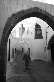 Image du Maroc Professionnelle de  Une femme marche dans une rue des Habous, la succession des balèvres, moulure, arches, portiques et autres éléments décoratifs, projetent les visiteurs dans l’ardeur avec un élan de force et de fierté d’une époque révolue. Conçu par les architectes Auguste Cadet et Edmond Brion d’après l’ébauche d’un premier plan-masse dû à Albert Laprade, le quartier fait partie des endroits les plus pittoresques de Casablanca, il fut La construction n’avait commencé qu’après la première guerre mondiale. Situé près du Palais Royale, ce joyau architectural fait partie du patrimoine architectural de la ville, Lundi 12 Février 2007. Au centre le minaret de La mosquée Ben Youssef. (Photo / Abdeljalil Bounhar)
